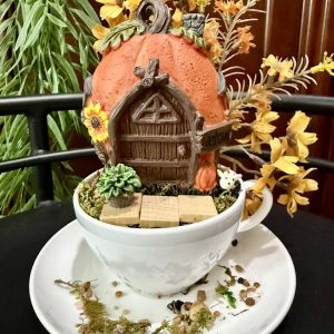 A pumpkin-shaped fairy house with a wooden door, sunflowers, and succulents sits atop a white teacup. Yellow-orange flowers and twigs decorate the scene, with scattered pebbles and moss on the saucer below.