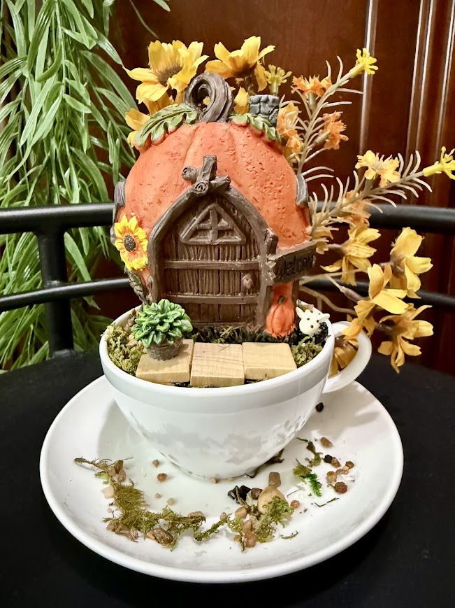 A pumpkin-shaped fairy house with a wooden door, sunflowers, and succulents sits atop a white teacup. Yellow-orange flowers and twigs decorate the scene, with scattered pebbles and moss on the saucer below.