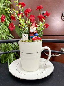 A tall white mug featuring a gnome with mushrooms and a small dog, surrounded by vibrant red flowers and green foliage.