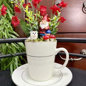 red-flower-gnome-dog-teacup-fairy-garden-decor A tall white mug featuring a gnome with mushrooms and a small dog, surrounded by vibrant red flowers and green foliage.