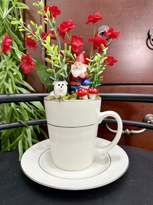 A tall white mug featuring a gnome with mushrooms and a small dog, surrounded by vibrant red flowers and green foliage.