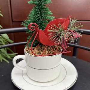 santas-sleigh-teacup-holiday-decor-christmas-centerpiece A red sleigh sits atop a white teacup filled with faux pebbles and a green pine tree, creating a festive Christmas scene.