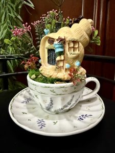 A shell-shaped fairy house with a tiny gnome outside is nestled in a white teacup with lavender and green floral patterns. The scene is surrounded by red flowers, greenery, and whimsical blue mushroom accents, all set on a matching saucer.