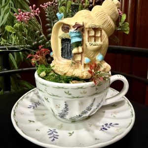 A shell-shaped fairy house with a tiny gnome outside is nestled in a white teacup with lavender and green floral patterns. The scene is surrounded by red flowers, greenery, and whimsical blue mushroom accents, all set on a matching saucer.