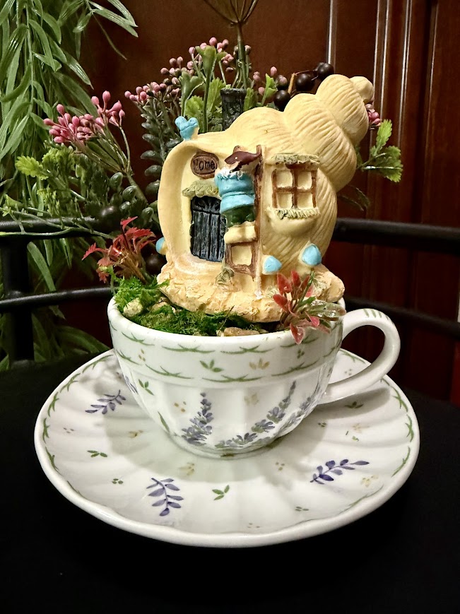 A shell-shaped fairy house with a tiny gnome outside is nestled in a white teacup with lavender and green floral patterns. The scene is surrounded by red flowers, greenery, and whimsical blue mushroom accents, all set on a matching saucer.