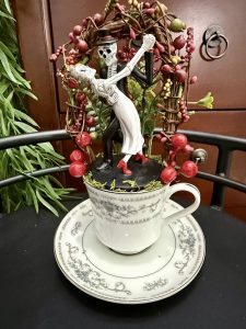 Spooky skeleton couple dancing under a rustic berry arch in a white and silver teacup, perfect for Halloween or Día de los Muertos décor.