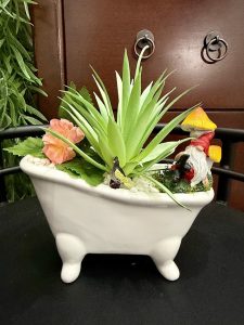 Whimsical bathtub-shaped planter filled with faux succulents, flowers, a gnome, butterfly, and miniature mushroom.