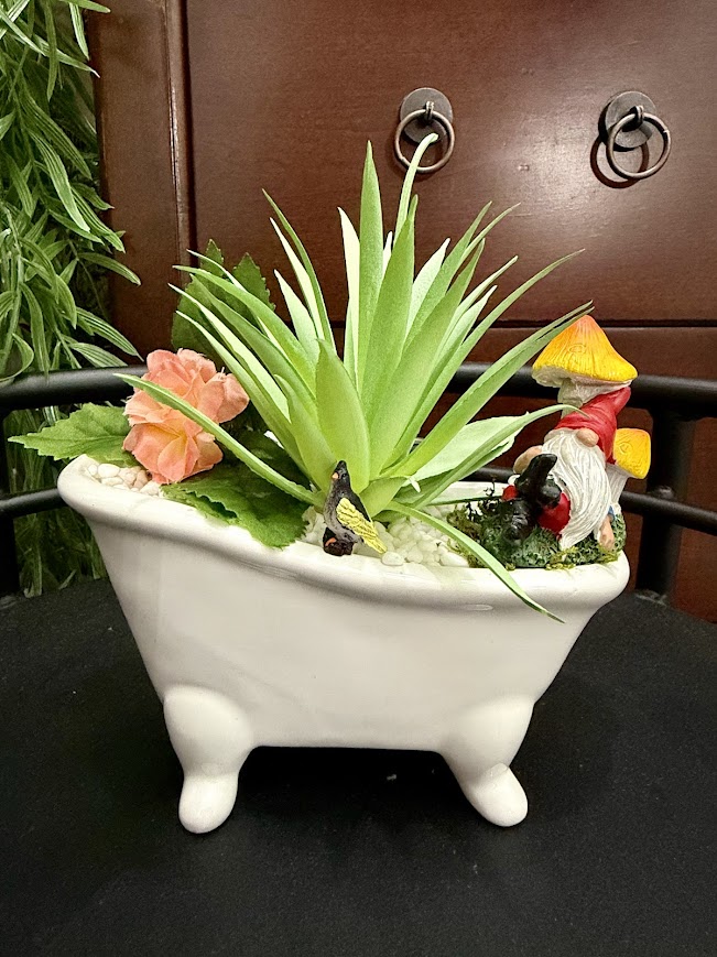 Whimsical bathtub-shaped planter filled with faux succulents, flowers, a gnome, butterfly, and miniature mushroom.