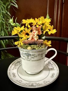 A fairy figurine with orange wings and a puppy sits on a wooden bench surrounded by vibrant yellow sunflowers in a delicate teacup with silver and gray floral designs on a matching saucer.