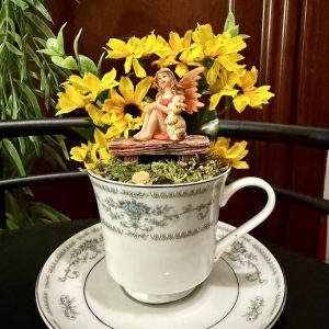 A fairy figurine with orange wings and a puppy sits on a wooden bench surrounded by vibrant yellow sunflowers in a delicate teacup with silver and gray floral designs on a matching saucer.
