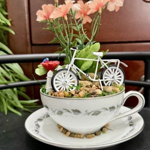 teacup-bike-flower-garden-pink-floral-miniature-decor A fine china teacup displays a miniature white bicycle resting on pebbles with soft pink flowers and greenery, evoking a peaceful countryside garden.