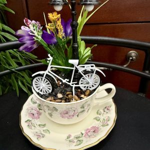 teacup-miniature-bicycle-lamppost-fairy-garden A vintage floral teacup holds a miniature white bicycle, faux flowers, pebbles, and a black lamppost, creating a whimsical garden street scene.