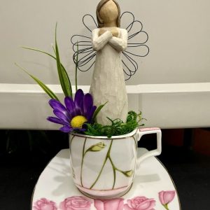 A square rose-patterned teacup holds a rustic angel figurine with wire wings, standing in a small moss garden accented with purple flowers.