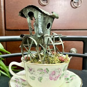 vintage-beer-cart-teacup-decor-rustic-fairy-garden A delicate rose-patterned teacup features a rustic miniature beer wagon with weathered wheels and mossy ground cover, blending old-world charm with fairy garden flair.
