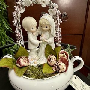 wedding-teacup-decor-bride-groom-miniature-centerpiece A white gravy boat is transformed into a romantic scene with a Precious Moments bride and groom figurine beneath a pink and white floral arch, surrounded by moss and faux roses.