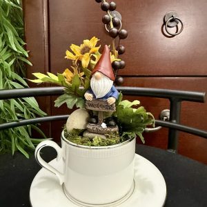 welcome-gnome-teacup-garden-centerpiece-decor A gnome holding a "Welcome" sign sits in a white teacup garden decorated with faux sunflowers, berries, moss, and stones.