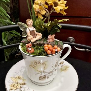 A floral teacup filled with a sitting fairy figurine, surrounded by faux yellow flowers, mushrooms, a squirrel, and a fox, creating a charming woodland scene.